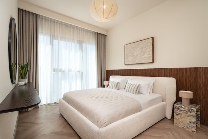 Dhoma gjumi elegante me shtroja prej pelushi, dekor modern dhe dritë të bollshme natyrore përmes dritareve të mëdha