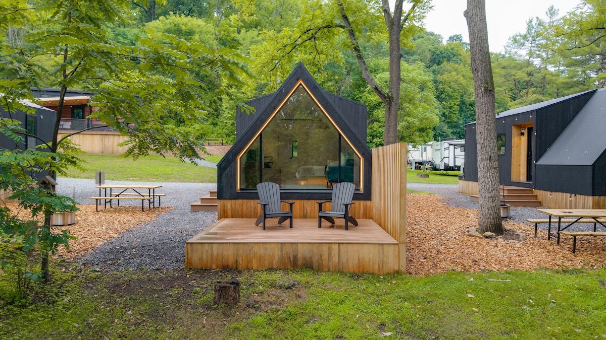 A tiny home exterior is showcased with a private patio featuring two Adirondack chairs. Natural wood decking extends out, surrounded by greenery and gravel paths. Nearby, picnic tables are set among a forested backdrop, enhancing the serene setting.