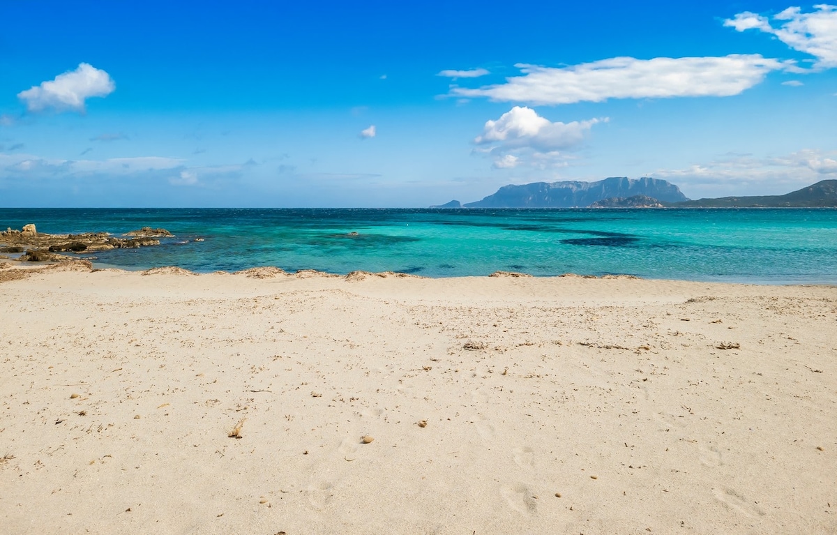 A sandy beach extends along a clear shoreline, where calm turquoise waters meet the coast. Gentle waves create a serene atmosphere, while distant mountains are visible against a bright blue sky dotted with a few clouds.