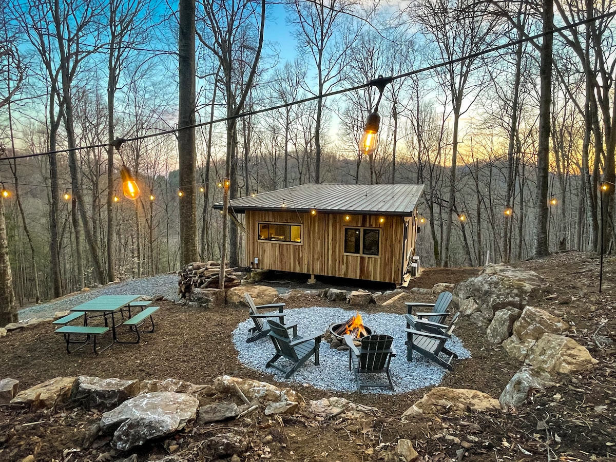 A modern cabin is positioned amidst trees, featuring a carefully arranged outdoor gathering space. Four Adirondack chairs surround a fire pit, illuminated by string lights overhead. Gravel pathways and picnic tables complement the natural landscape, enhancing a sense of tranquility and connection with the surroundings.