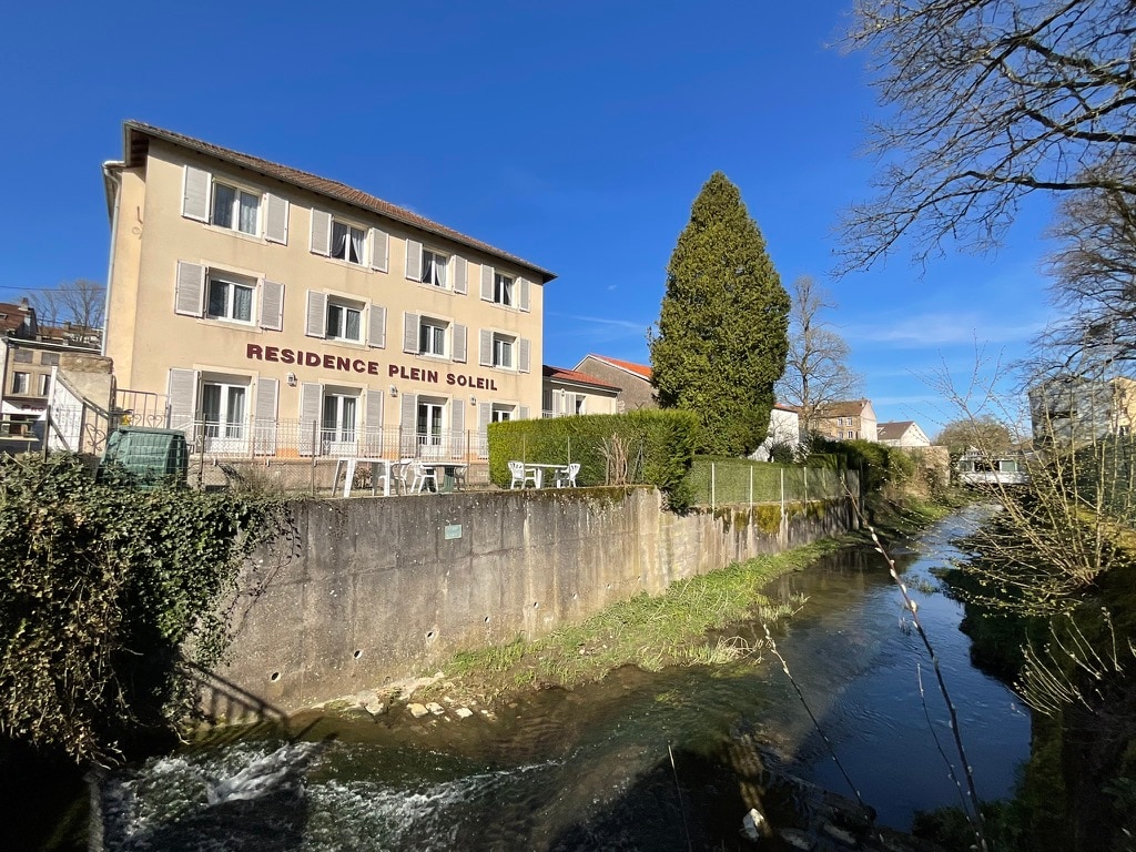 The image captures the exterior of the residence, featuring a three-story building with a sunny façade and large windows. A lush tree and grassy area are visible in front, alongside a gently flowing stream that runs beside the property. Outdoor seating is present for guest use.
