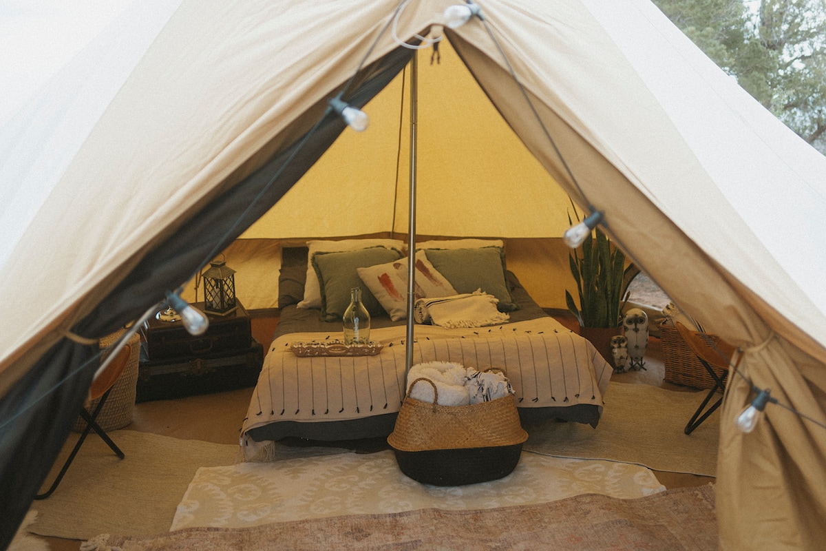An inviting glamping tent interior is displayed, featuring a spacious sleeping area with a neatly made bed, accented by soft pillows and blankets. Warm lighting is created with decorative string lights. Decorative rugs and baskets add a cozy touch to the surroundings.