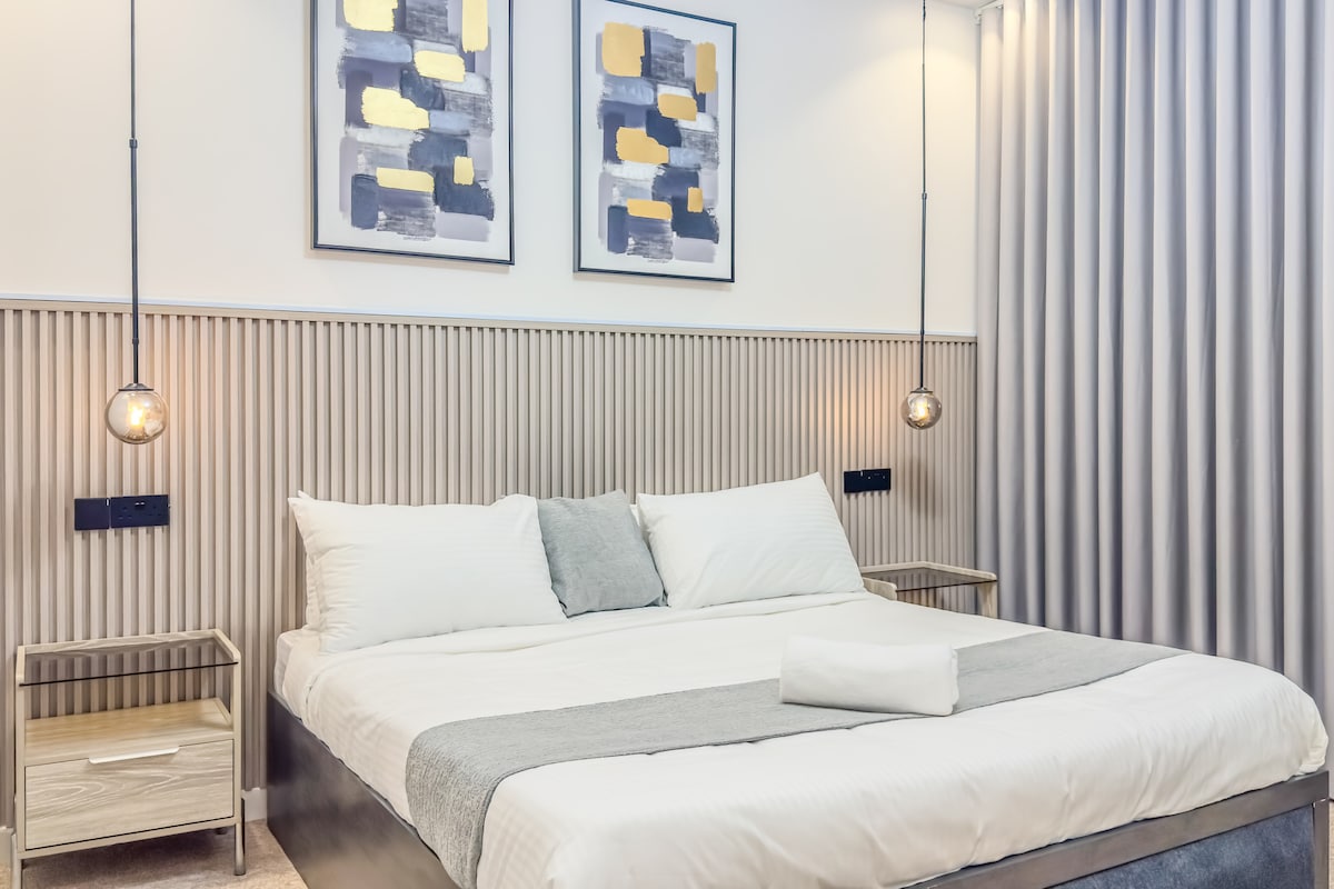 The master bedroom features a spacious bed topped with premium bedding. Twin bedside tables hold modern lamps, while stylish artwork adorns the walls above. Soft lighting is provided by pendant fixtures, complemented by textured curtains that create a soothing ambiance.