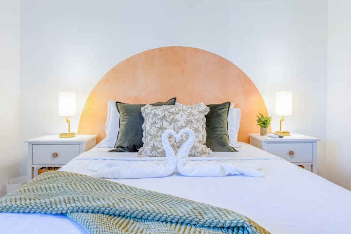 חדר שינה קווין מקסים עם עיצוב בוהו - שיק, מודגש על ידי קיר מבטא עם גוון אפרסק, כריות מפוארות ומצעים רכים בשכבות. שידות לילה תואמות עם מנורות קריאה וזריקה נעימה יוצרים את האווירה המושלמת לרגיעה ונוחות.