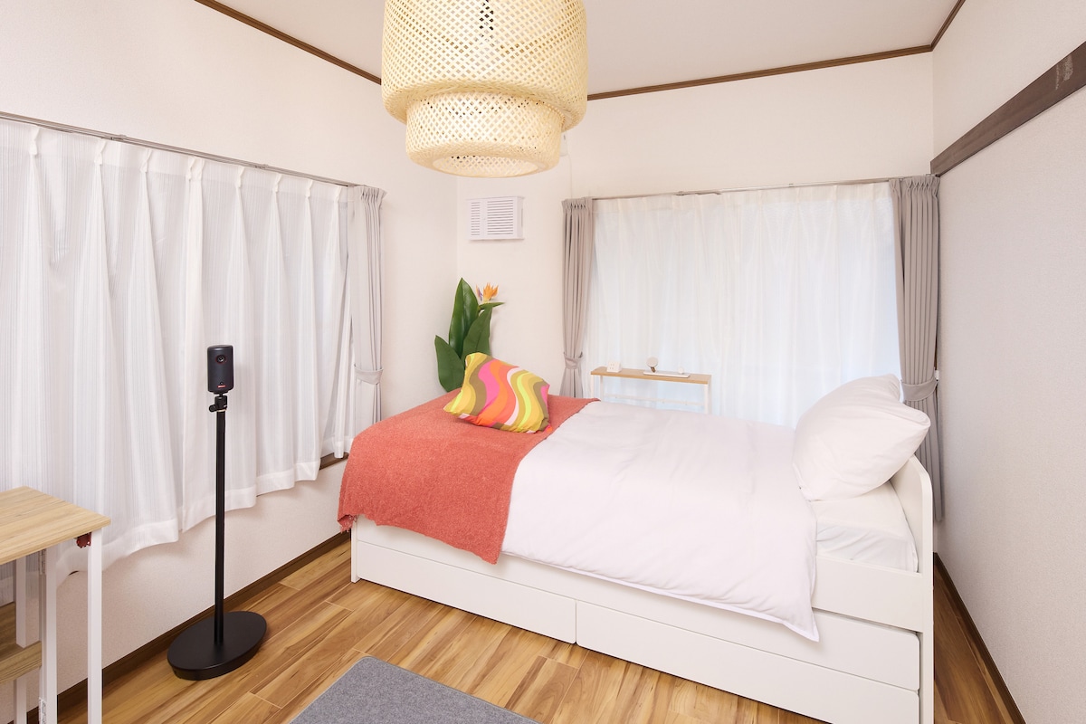 A compact room features a spacious bed with a bright orange throw and colorful cushions, complemented by natural light from sheer curtains. A potted plant adds a touch of greenery, while a small wooden table stands nearby, completing the functional and inviting space.