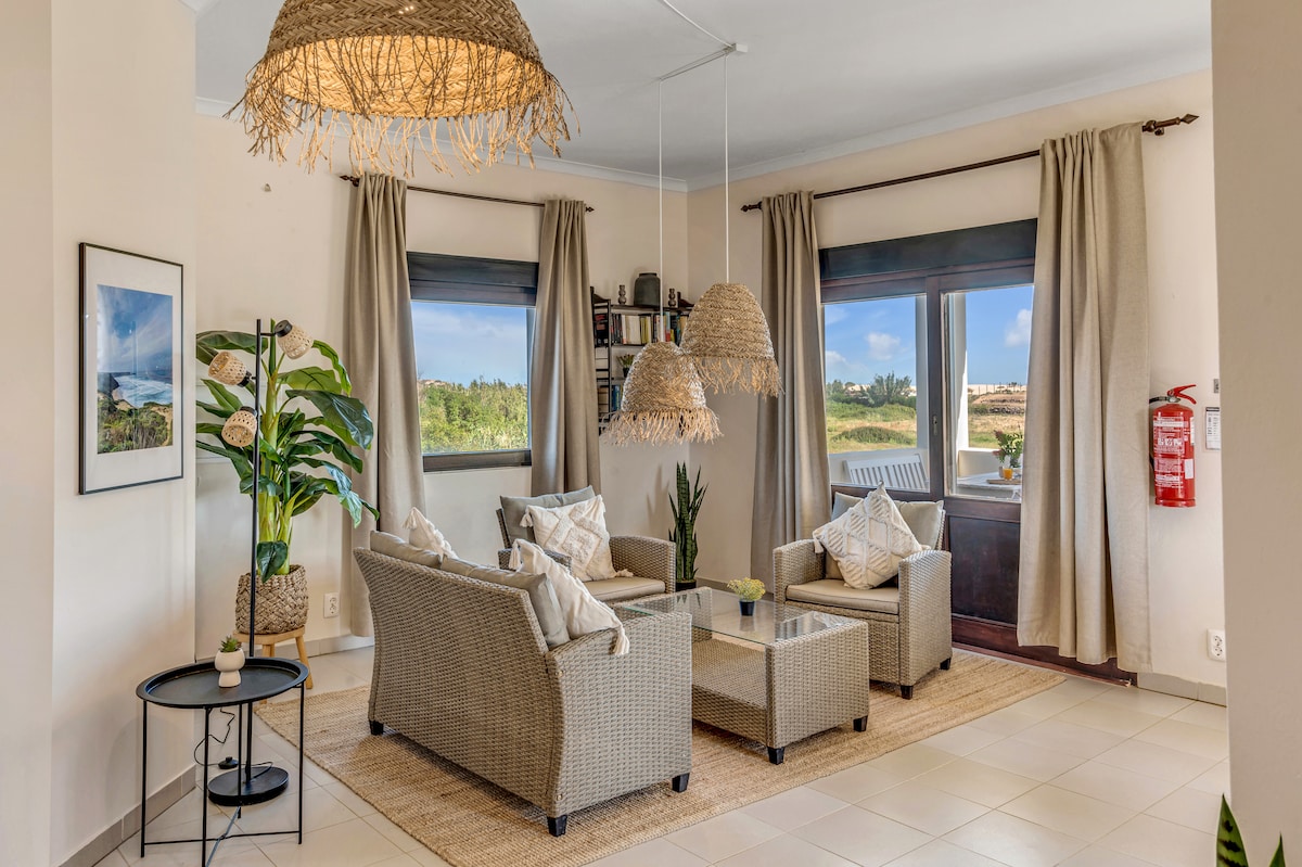 A welcoming living area features comfortable seating arrangements with natural fiber accents. Large windows provide views of the surrounding landscape, while indoor plants add a touch of greenery. Soft lighting is provided by woven pendant lamps, enhancing the room's bright and airy feel.