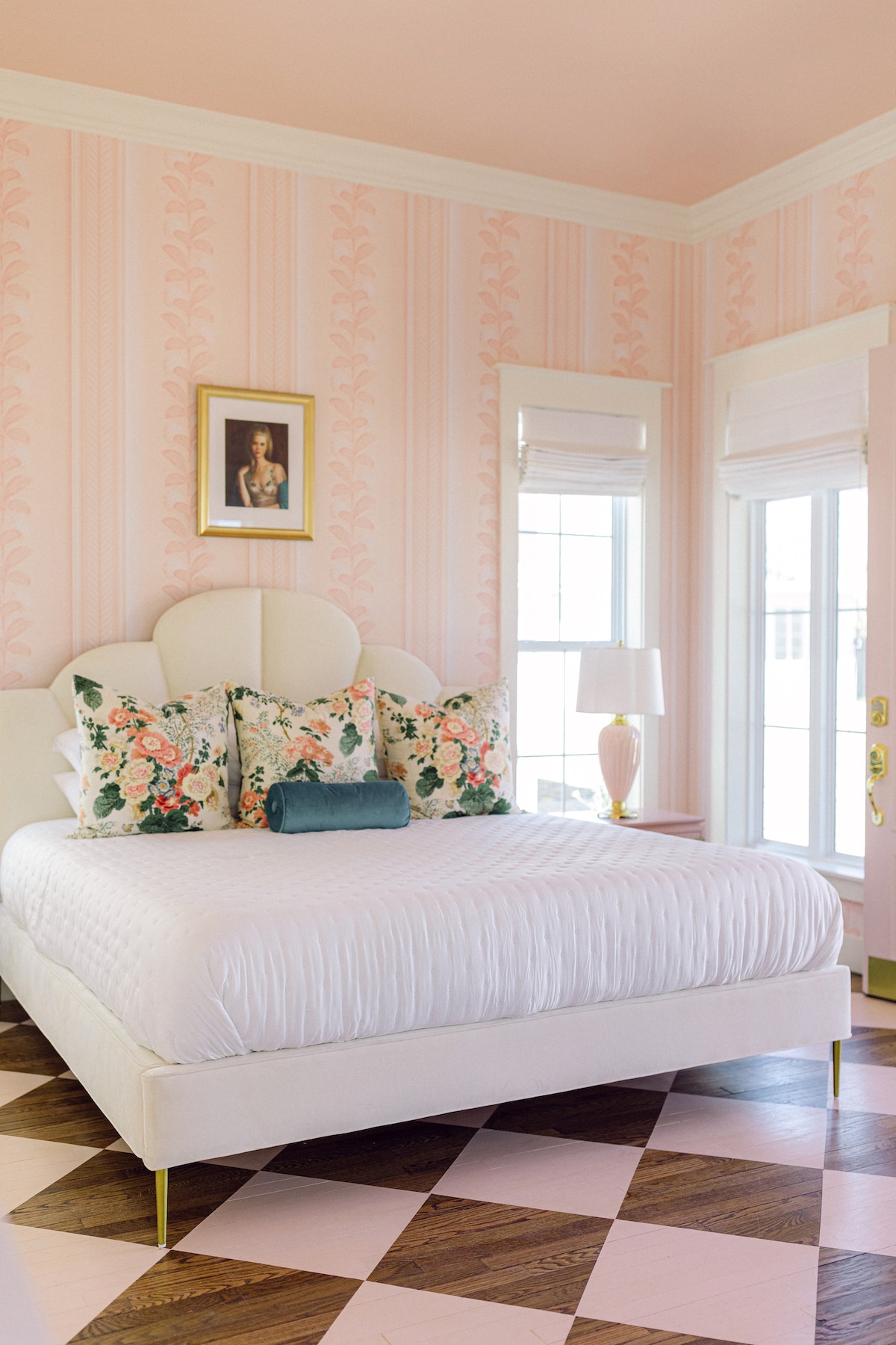 A softly decorated bedroom features a spacious white bed adorned with floral accent pillows. Light pink wallpaper with subtle patterns covers the walls. Two large windows allow natural light to enter, complemented by elegant window treatments and a table lamp. The floor showcases a classic checkerboard design.