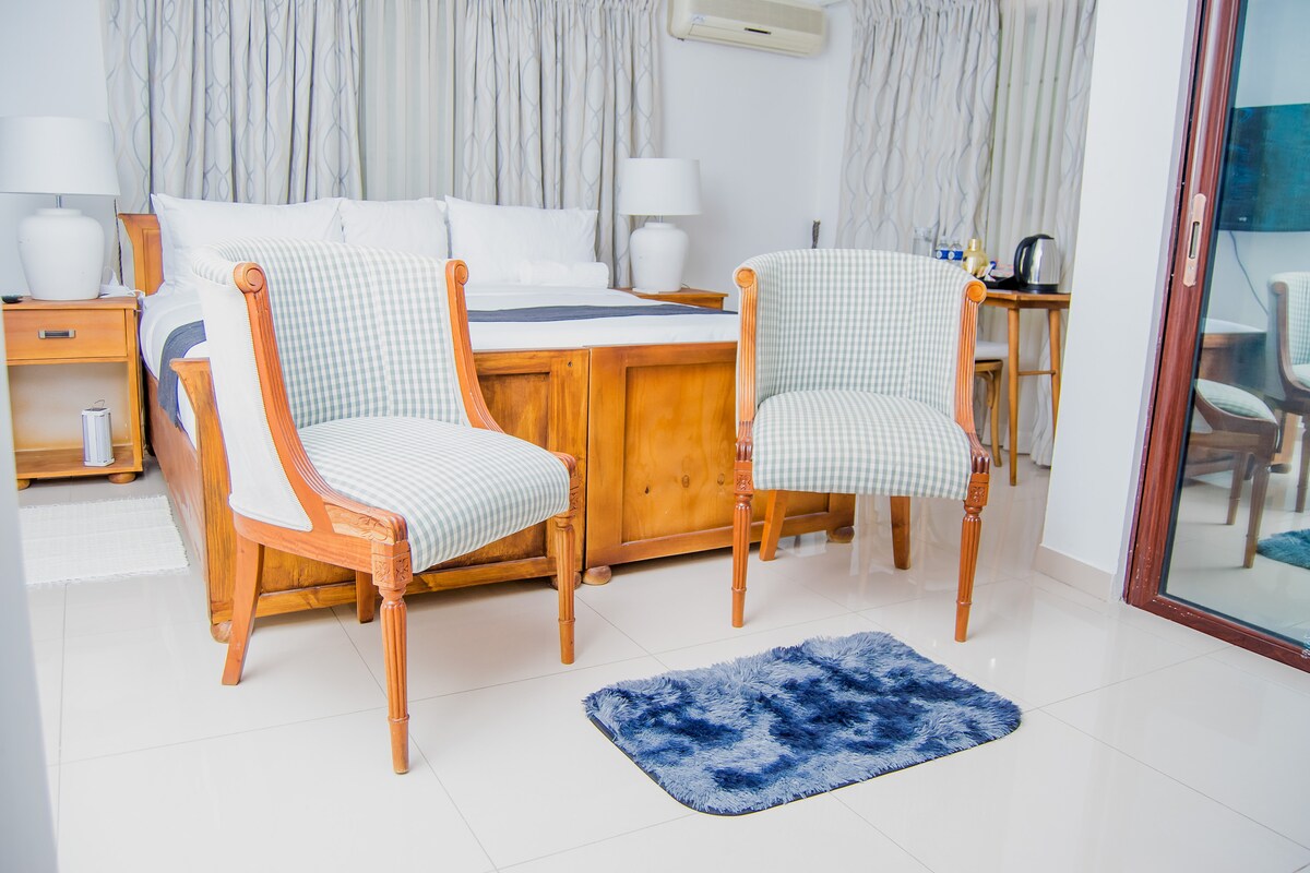 A cozy bedroom is featured with a spacious bed adorned with crisp linens. Two patterned chairs are positioned at the foot of the bed, facing a plush blue rug. Soft natural light enters through large windows, enhancing the serene atmosphere.