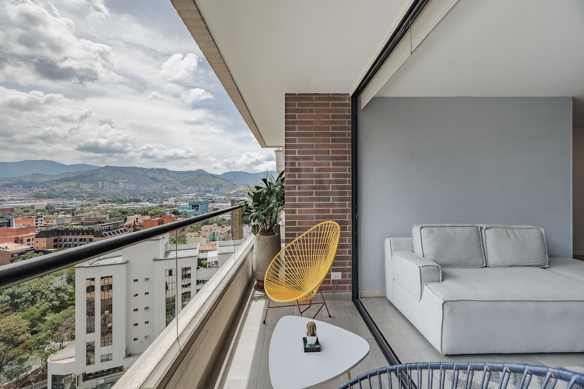 A spacious balcony area features a light gray sofa and a yellow chair. Potted plants are positioned nearby, with panoramic views of the city and surrounding mountains visible through large glass sliding doors. Natural light fills the space, creating an inviting atmosphere.