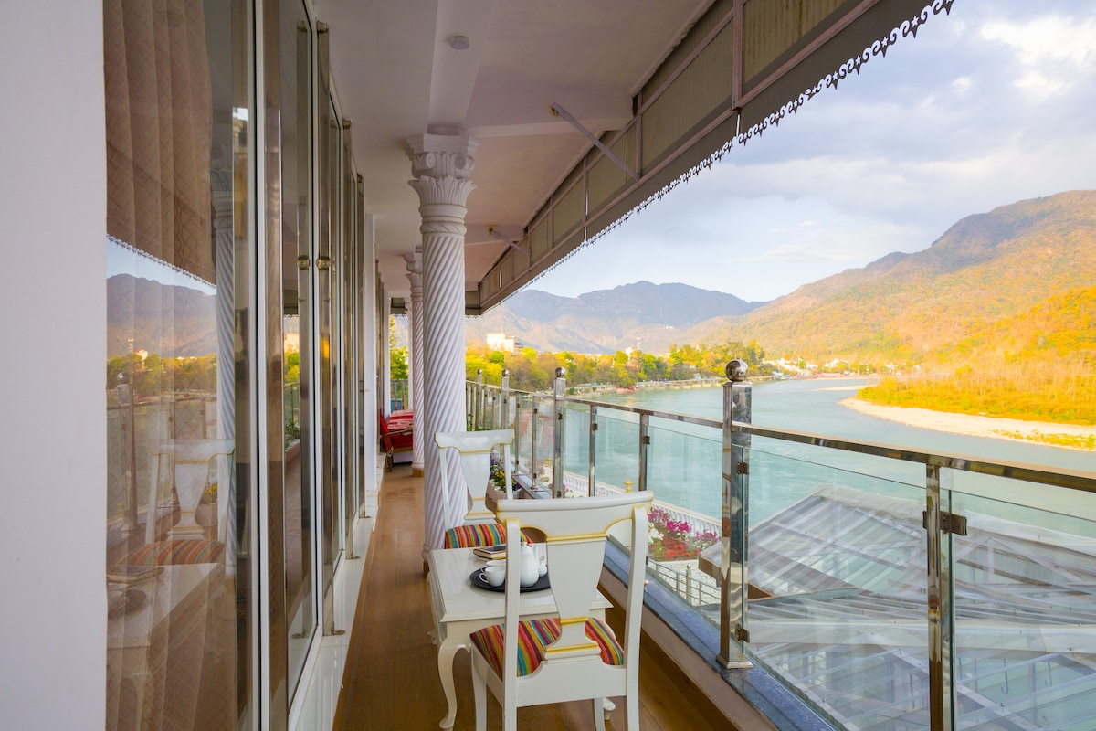 A spacious balcony features a glass railing with scenic views of the River Ganges and surrounding mountains. Brightly colored striped chairs and a small table provide a comfortable spot for relaxation. The soft morning light filters through large floor-to-ceiling windows, enhancing the tranquil ambience.