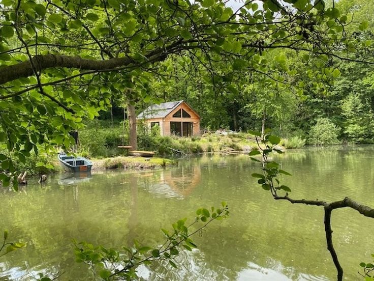 A wooden cabin is visible beside a calm, reflective pond, surrounded by lush greenery. The structure features large windows, allowing natural light to enter. A small boat rests gently on the water, adding to the serene setting within a natural park environment.