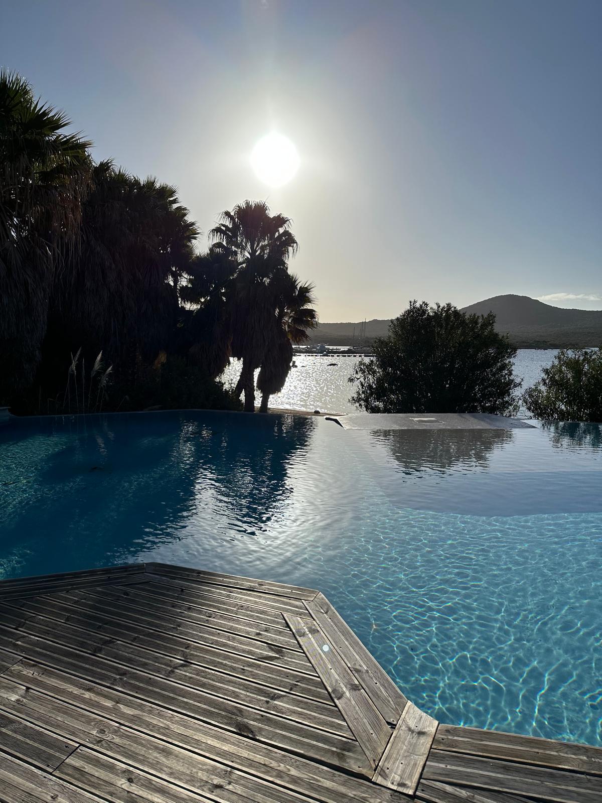 A serene pool area is highlighted under a radiant sun, surrounded by lush palm trees. The pool's calm water reflects the sky, creating a tranquil scene that connects with the natural landscape in the background.