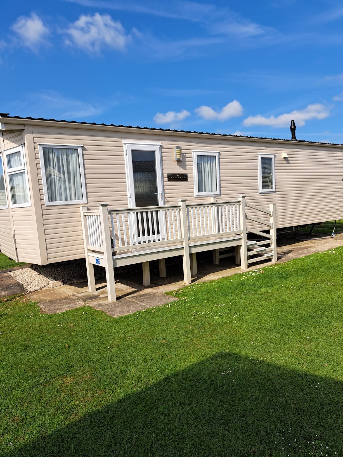 The exterior of the static caravan is shown, featuring a light-colored façade with white trim. A small deck is visible at the entrance, accompanied by steps leading down to the green lawn. Clear blue skies and a few clouds create a bright atmosphere.