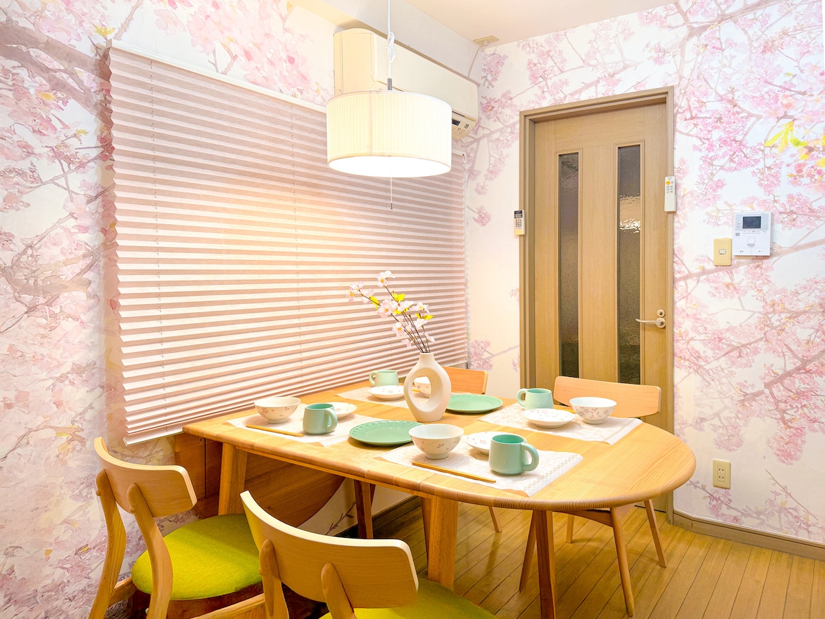 A dining area features a wooden table surrounded by green accent chairs. The space is decorated with cherry blossom wallpaper and is softly illuminated by a ceiling light. Simple white dinnerware is set on the table, accompanied by a decorative vase with flowers.