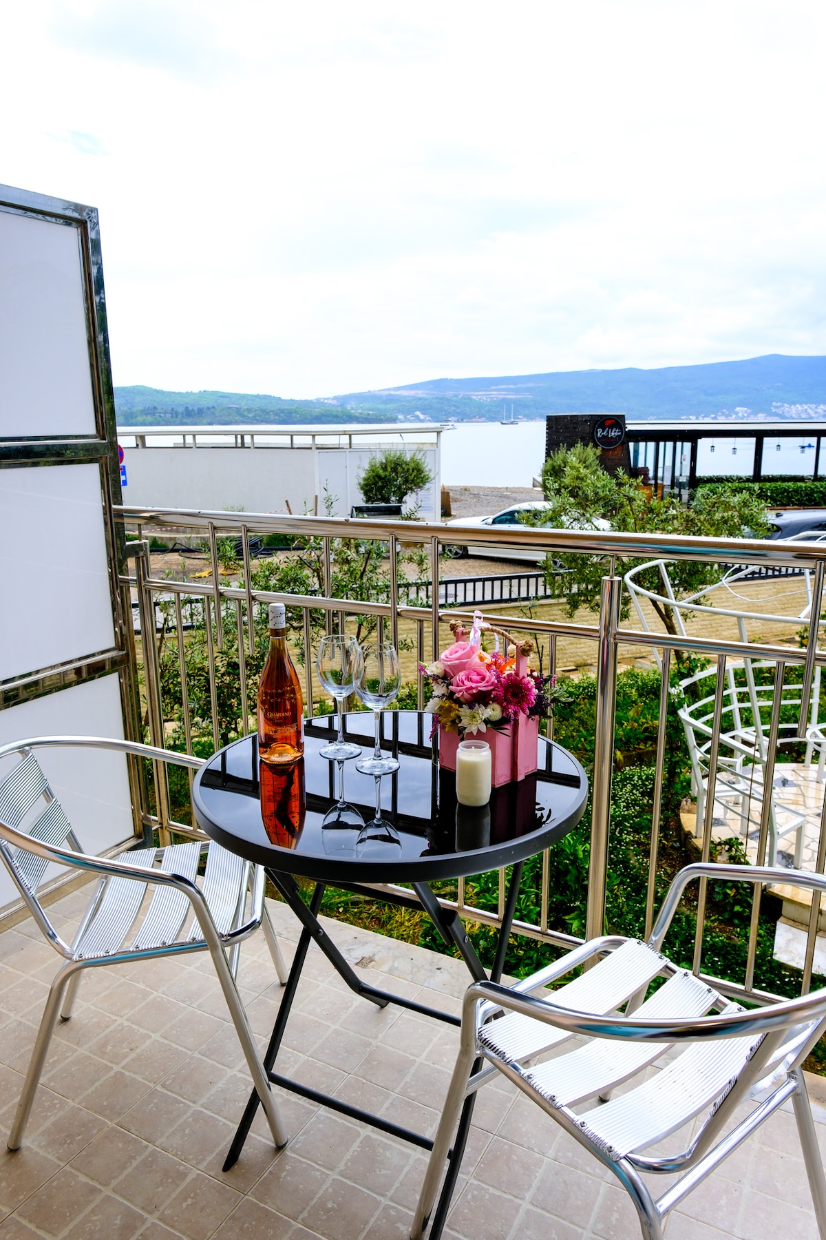 A balcony view features a circular table set with two chairs. A bottle with glasses and a small floral arrangement is placed on the table. The backdrop includes a scenic vista of water and greenery, framed by modern railing and nearby foliage.