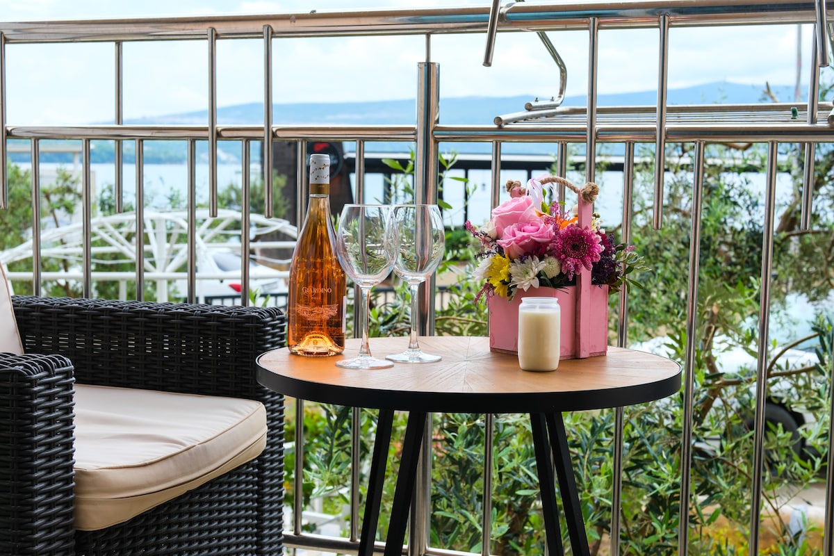 A small outdoor table is set with two glasses, a bottle of rosé wine, and a decorative arrangement of flowers in a pink container. A candle is placed on the table, and a wicker chair is positioned beside it, framed by greenery with a view of the water in the background.