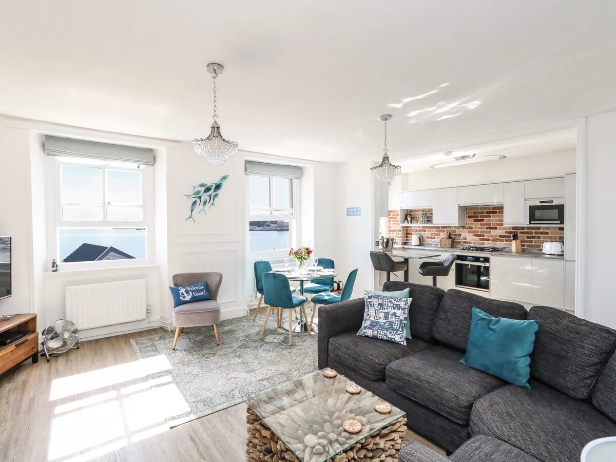 The spacious lounge area features a comfortable dark sofa with decorative cushions, a glass coffee table, and a light rug. An open-plan kitchen with modern appliances is visible, alongside a dining table with four chairs. Seamless natural light enters through large windows, offering views of the bay.