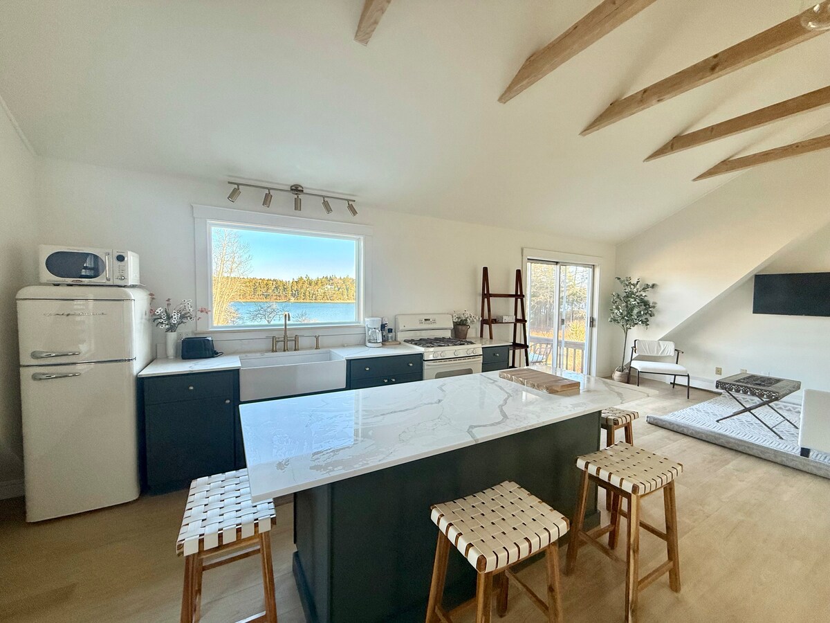 An airy kitchen features a large island with seating made of woven materials. Natural light streams through a wide window, offering views of the surrounding landscape. Modern appliances, including a vintage-style refrigerator, are present, complemented by wooden beams overhead.