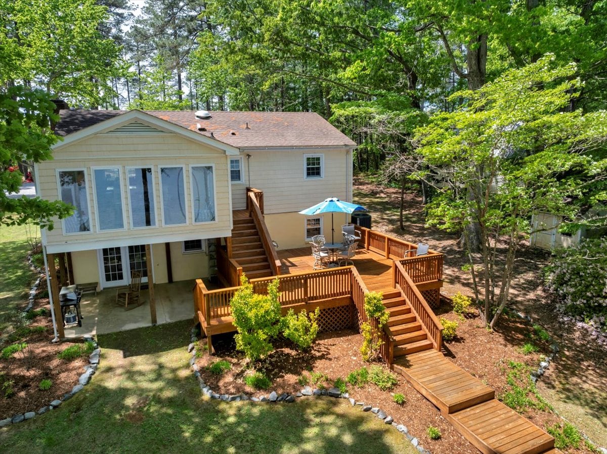 An elevated view of a split-level house showcases a spacious deck surrounded by lush greenery. Comfortable outdoor seating is visible under a blue umbrella, with wooden steps leading down to the yard. A gas grill is positioned on the patio, enhancing the outdoor experience.