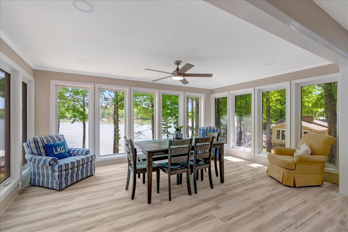 A sunlit dining area features a wooden table surrounded by black chairs, complemented by two comfortable chairs near the windows. Large windows provide panoramic views of the lake, while a ceiling fan adds a touch of comfort.