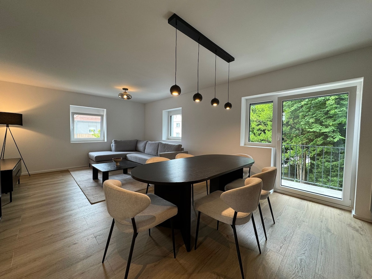 A spacious living area is showcased, featuring a large dark table surrounded by light-colored chairs. A comfortable gray sofa is positioned along one wall, while large windows allow natural light to fill the room. A floor lamp stands nearby, adding to the overall ambiance.