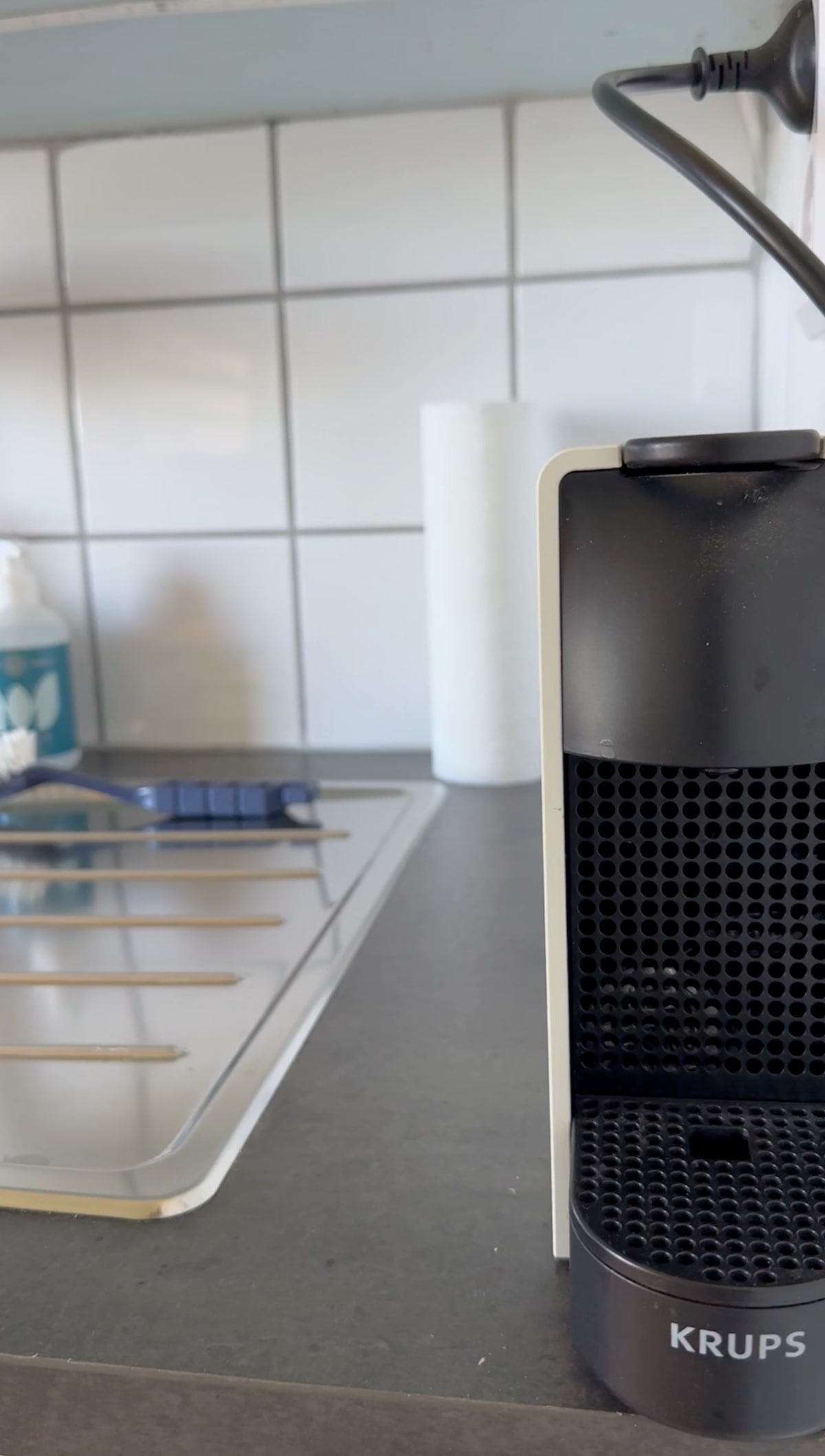 A coffee machine is positioned on a countertop, showcasing a sleek black and white design. The background features a stainless-steel stove with visible cooking utensils, a roll of paper towels, and cleaning supplies, all against a tiled wall.