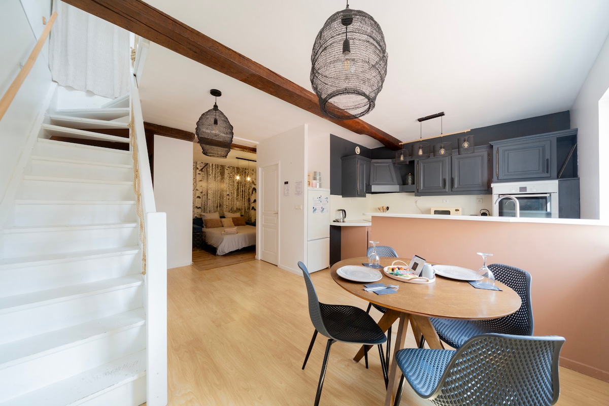 A spacious dining area features a round wooden table set for a meal, accompanied by four modern chairs. Two hanging pendant lights add a unique touch. The open layout reveals a glimpse of the kitchen and leads to a white staircase, enhancing the inviting atmosphere.