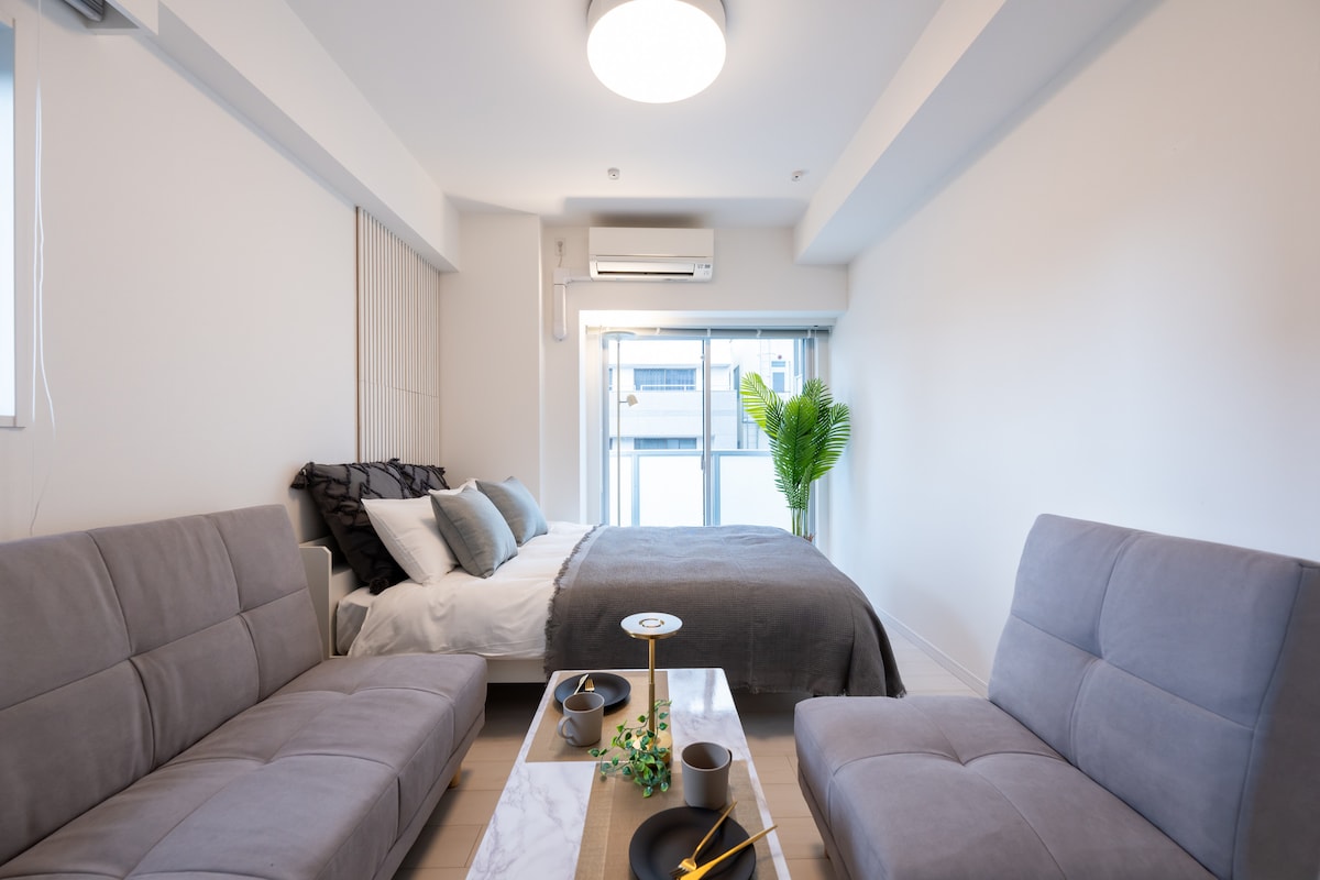 A well-lit room features a double bed with gray bedding and several cushions. Two gray sofas are positioned across from a small marble table. A plant is located near the window, allowing natural light to fill the space. Air conditioning is mounted on the wall.