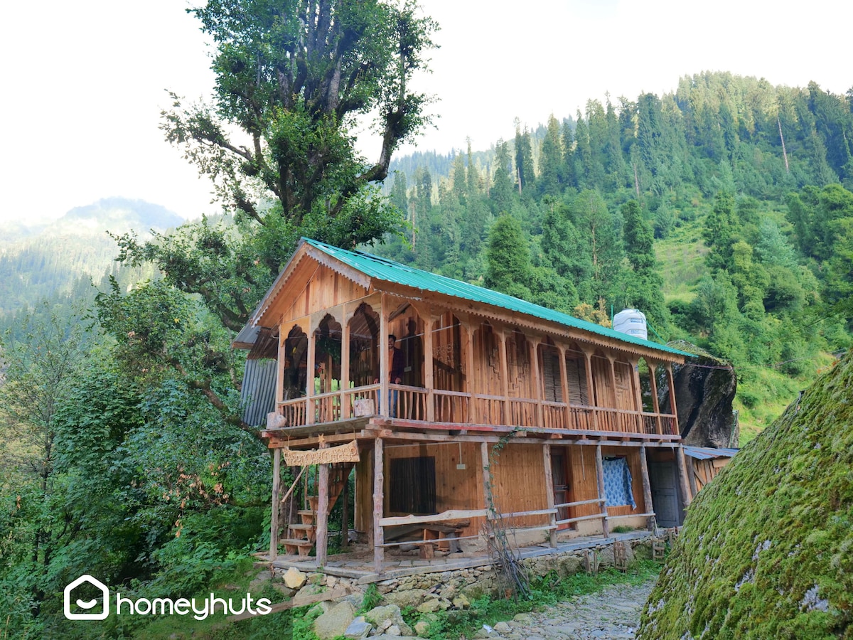A wooden structure is nestled among lush green trees, showcasing a multi-level design with a sloped roof. A balcony extends from the upper level, offering views of the surrounding mountains. Natural elements integrate harmoniously with the outdoor setting, providing a peaceful ambiance.
