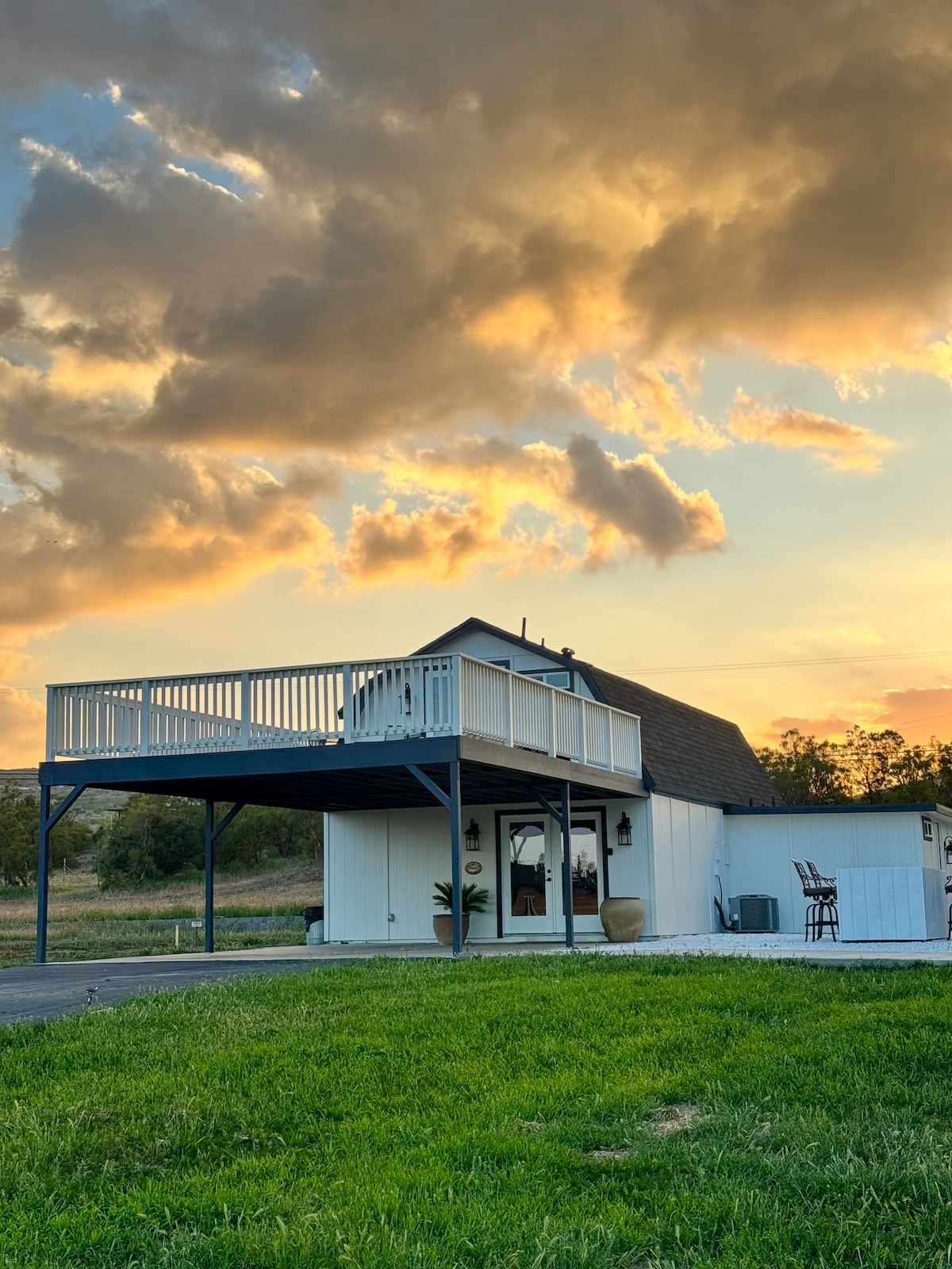 A two-story barndominium is showcased against a dramatic sunset, with hues of orange and yellow reflecting in the sky. A spacious balcony with chairs is visible on the upper level, while a green lawn surrounds the structure.