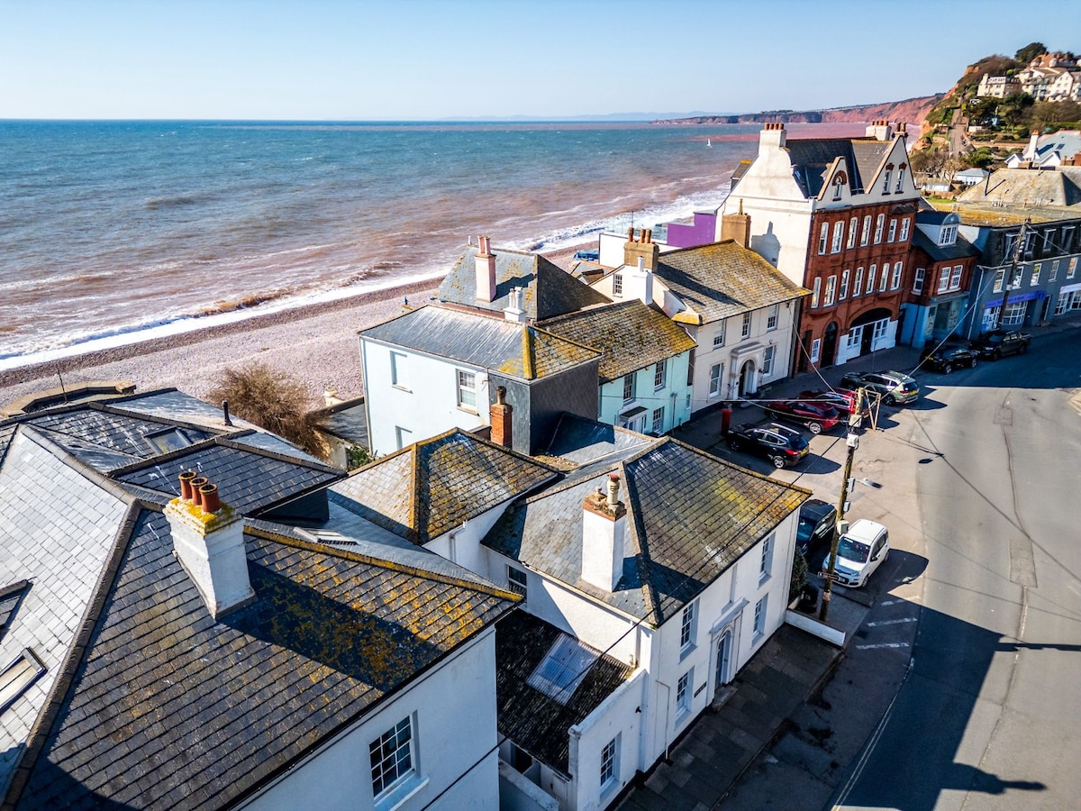 Aerial view reveals colorful cottages positioned along a coastal road, with the expansive beach and ocean in the background. The charming architecture and the proximity to the sea highlight the unique character of the area, while the peaceful shoreline invites exploration.