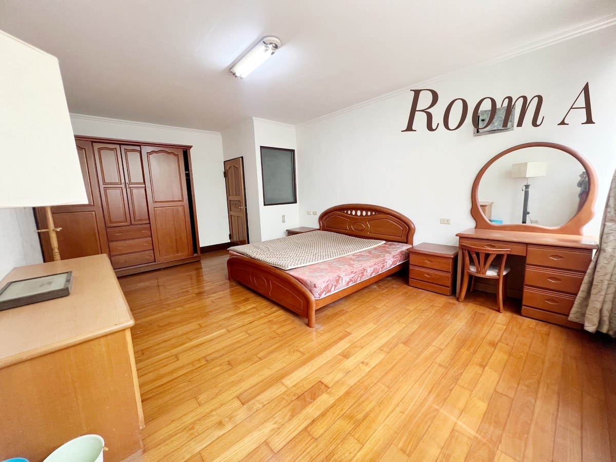 A spacious room is featured, containing a large bed and wooden furniture including a dresser with a round mirror and a desk. A wardrobe is positioned against one wall. The wooden flooring adds warmth, while natural light brightens the area.