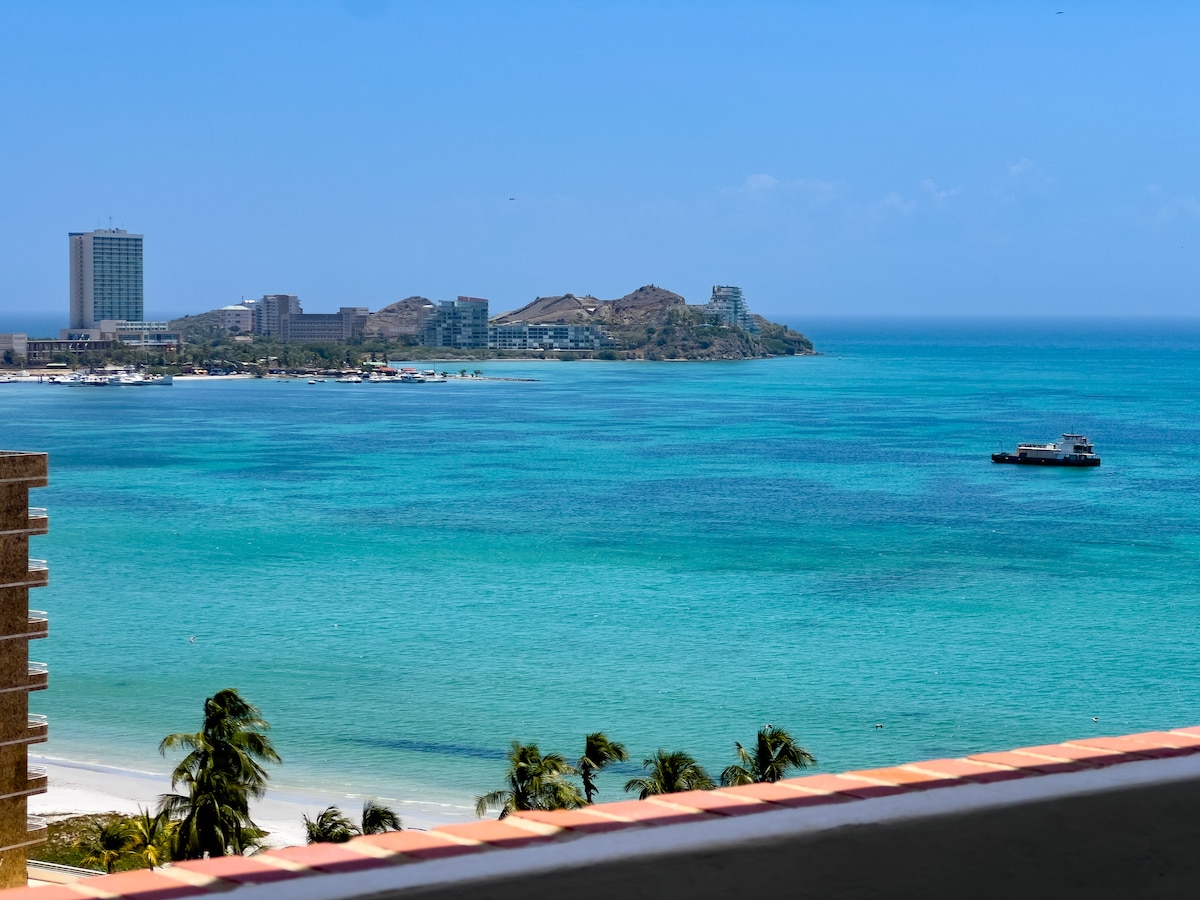 A serene view of the turquoise sea stretching towards the horizon, with soft waves gently lapping at the shore. A distant shoreline reveals a mix of modern buildings and greenery, while a small boat can be seen gliding across the water, enhancing the coastal scene.