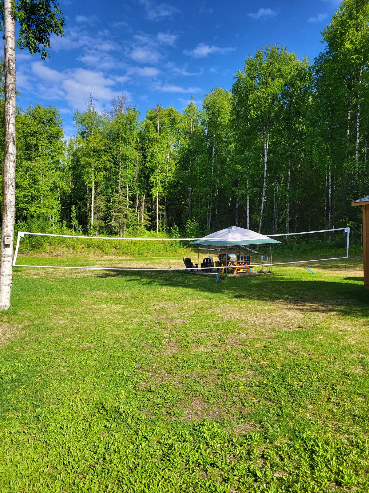 A well-maintained outdoor space features a volleyball net stretching across a grassy area, surrounded by tall trees. A tent with multiple chairs is set up nearby, providing a sheltered spot for relaxation. The bright sky and scattered clouds add to the inviting landscape.