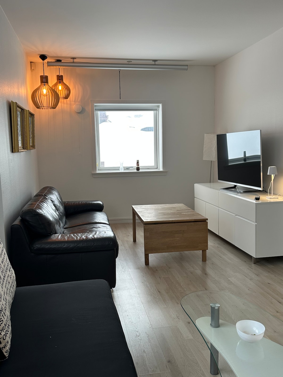 A comfortable living area is furnished with a dark leather sofa and a light wooden coffee table. A modern television is positioned on a white media console. Natural light enters through a window, complemented by decorative pendant lighting and a floor lamp.