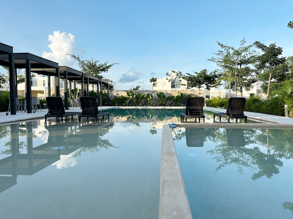 The swimming pool is surrounded by lush greenery and features a modern design. Several black lounge chairs are positioned on the pool deck, providing relaxation spots under the open sky. The calm water reflects the structures and trees nearby, emphasizing a serene environment.
