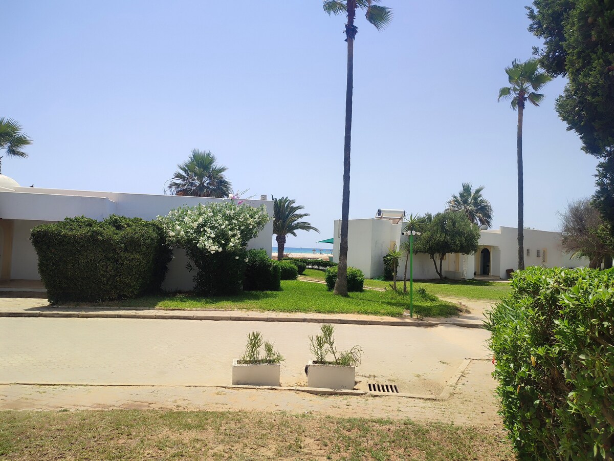 A peaceful outdoor area is shown, featuring palm trees and well-maintained greenery. White bungalow structures are visible in the background, while a glimpse of the swimming pool can be seen beyond the landscaping. The pathway is bordered by low shrubs and flowering plants.