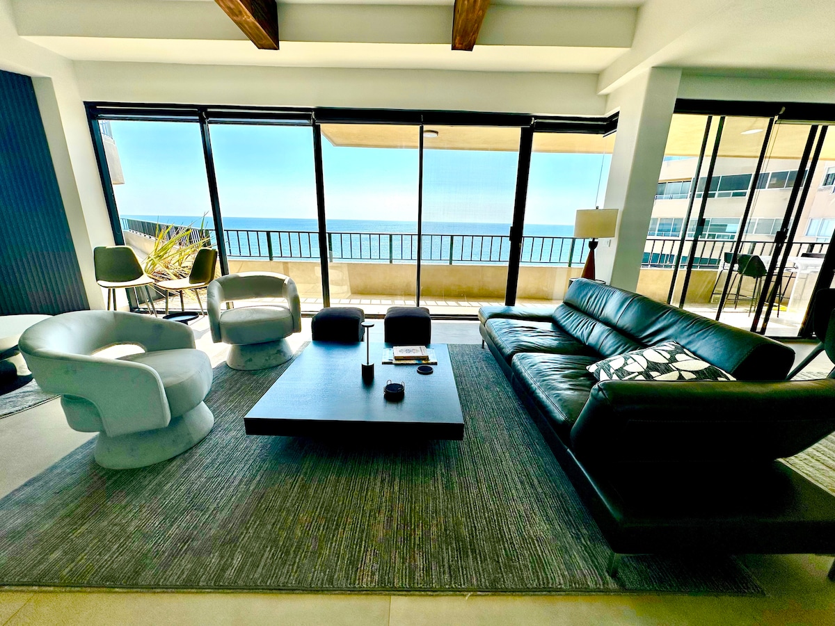 The open concept living area features floor-to-ceiling windows showcasing ocean views. A sleek black sofa is centered on a modern area rug, complemented by white armchairs. A coffee table sits centrally, while natural light floods the space, creating a serene atmosphere.