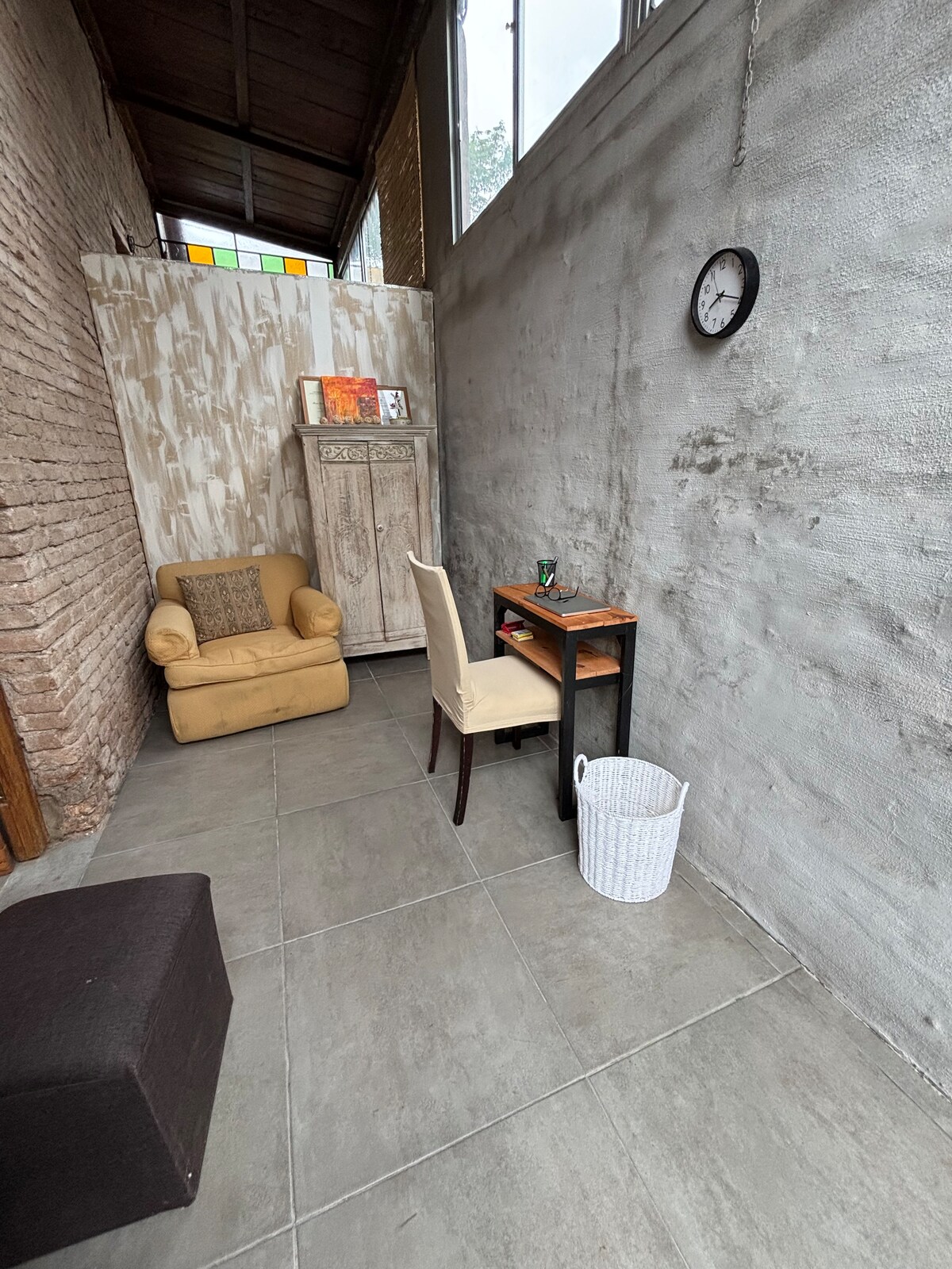 A small, well-lit corner features a beige armchair next to a wooden desk. A white clock is mounted on the textured gray wall, and a decorative cabinet stands nearby. Natural light streams in through the high windows, illuminating the tiled floor.