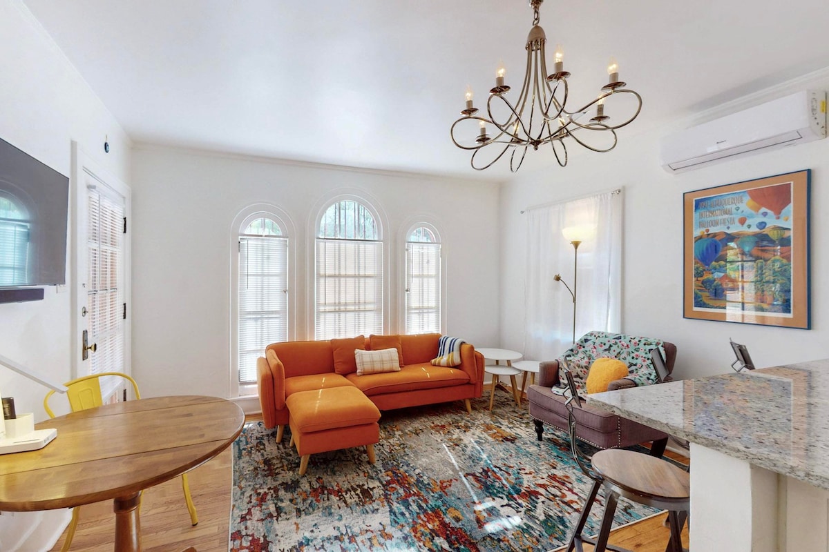 A living area features large arched windows allowing natural light to fill the space. An orange sofa provides seating alongside a patterned area rug. A round dining table is positioned with a cozy armchair nearby. Decorative art hangs on the wall, complementing the room's ambiance.
