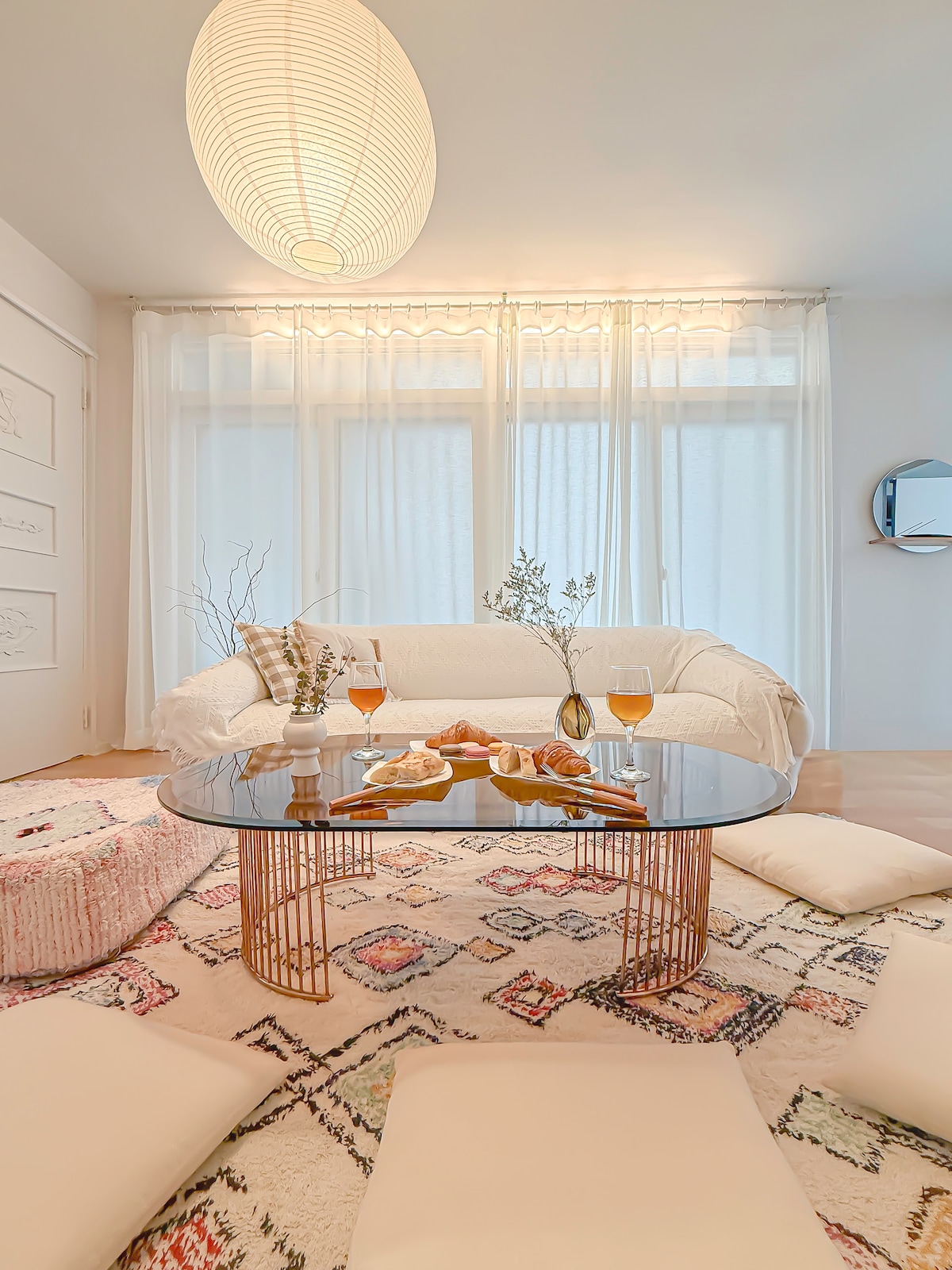 A light-filled living area features a low table set with food and drinks, surrounded by cushions on a patterned rug. Sheer curtains filter sunlight, creating a soft ambiance. A cozy sofa invites relaxation, complemented by a decorative wall mirror and pendant light overhead.