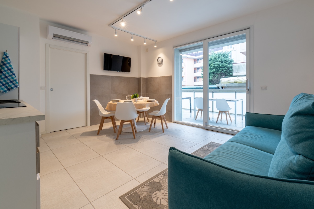 A modern living area is depicted, featuring a comfortable sofa in a soft hue. A dining table with white chairs accommodates a centerpiece. Large sliding glass doors allow natural light and offer views of the balcony, enhancing the space's openness.