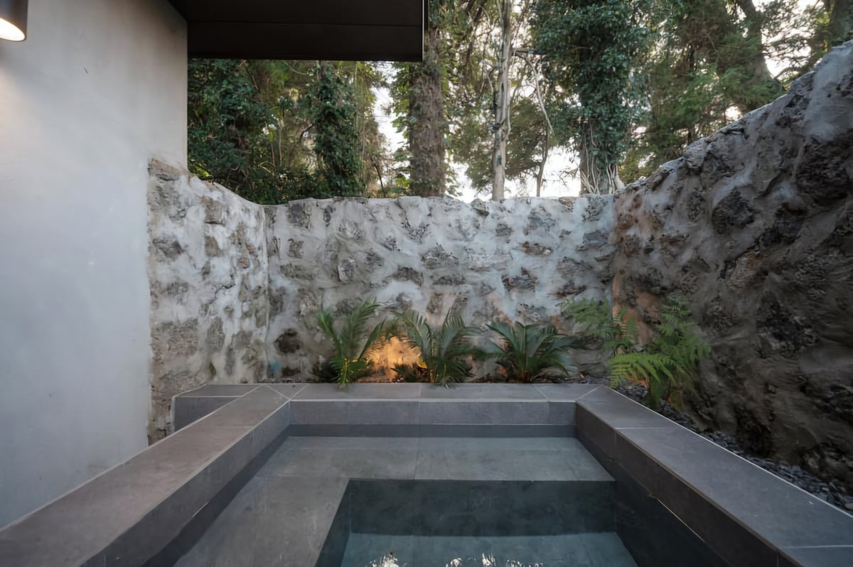 A serene outdoor hot tub is framed by a textured stone wall and lush greenery. The calm water reflects the surrounding foliage, creating a peaceful retreat. Soft lighting enhances the natural ambiance of the space, inviting relaxation and connection with nature.