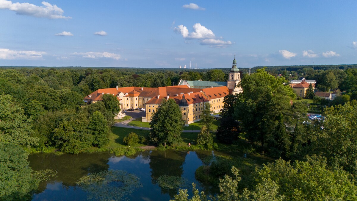 An expansive view showcases the historic buildings of a monastic complex surrounded by lush greenery. A serene pond reflects the structures and the clear blue sky, while trees add to the peaceful atmosphere of the landscaped park.
