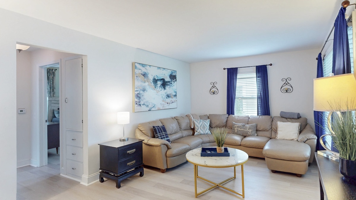 A comfortable living room features a cozy sectional sofa and a round, light-colored coffee table. Natural light streams through the windows adorned with navy curtains. A piece of wall art adds a touch of color to the neutral walls, contributing to a calm and inviting environment.