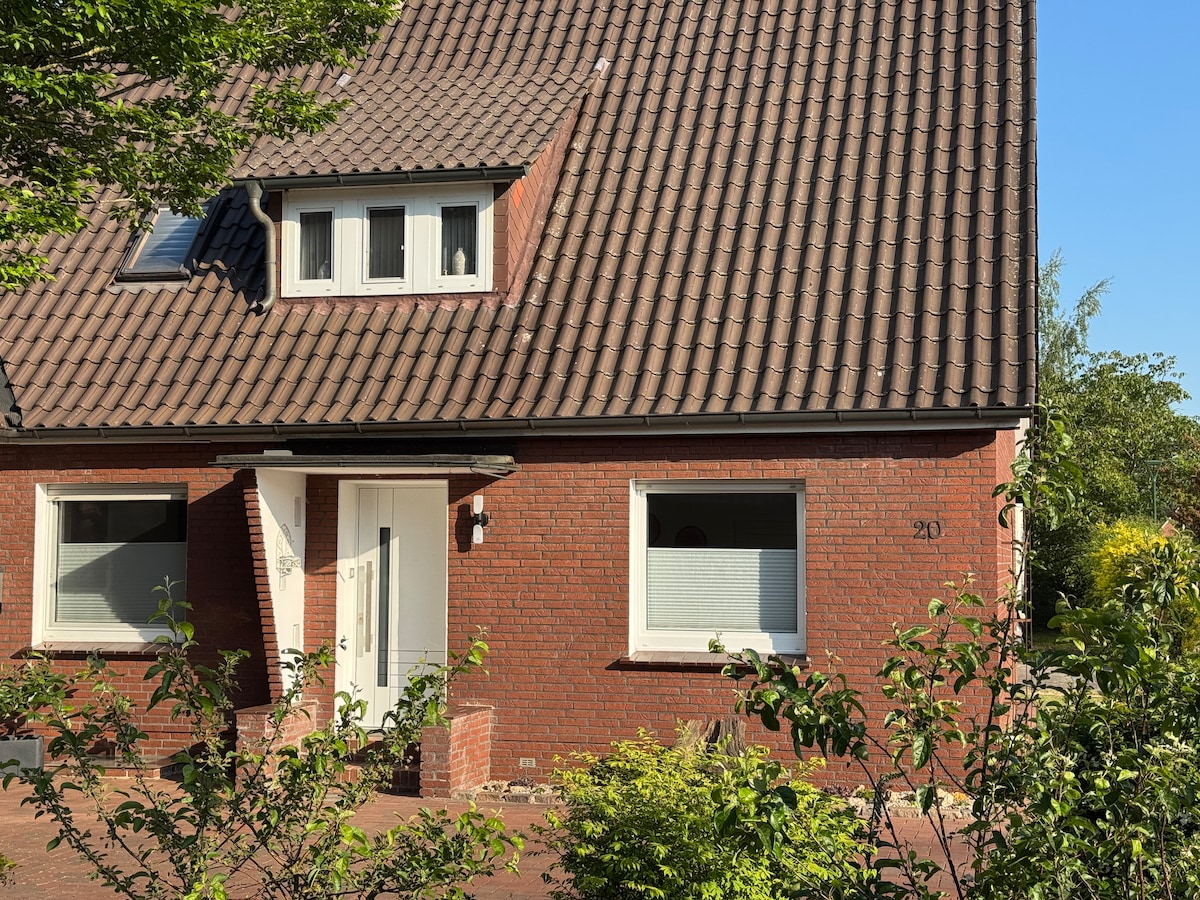 The exterior of the house features a red brick façade with a pitched roof covered in dark tiles. Two windows are positioned symmetrically on the first floor, providing natural light. A welcoming entrance door is framed by a small porch, and greenery surrounds the property.