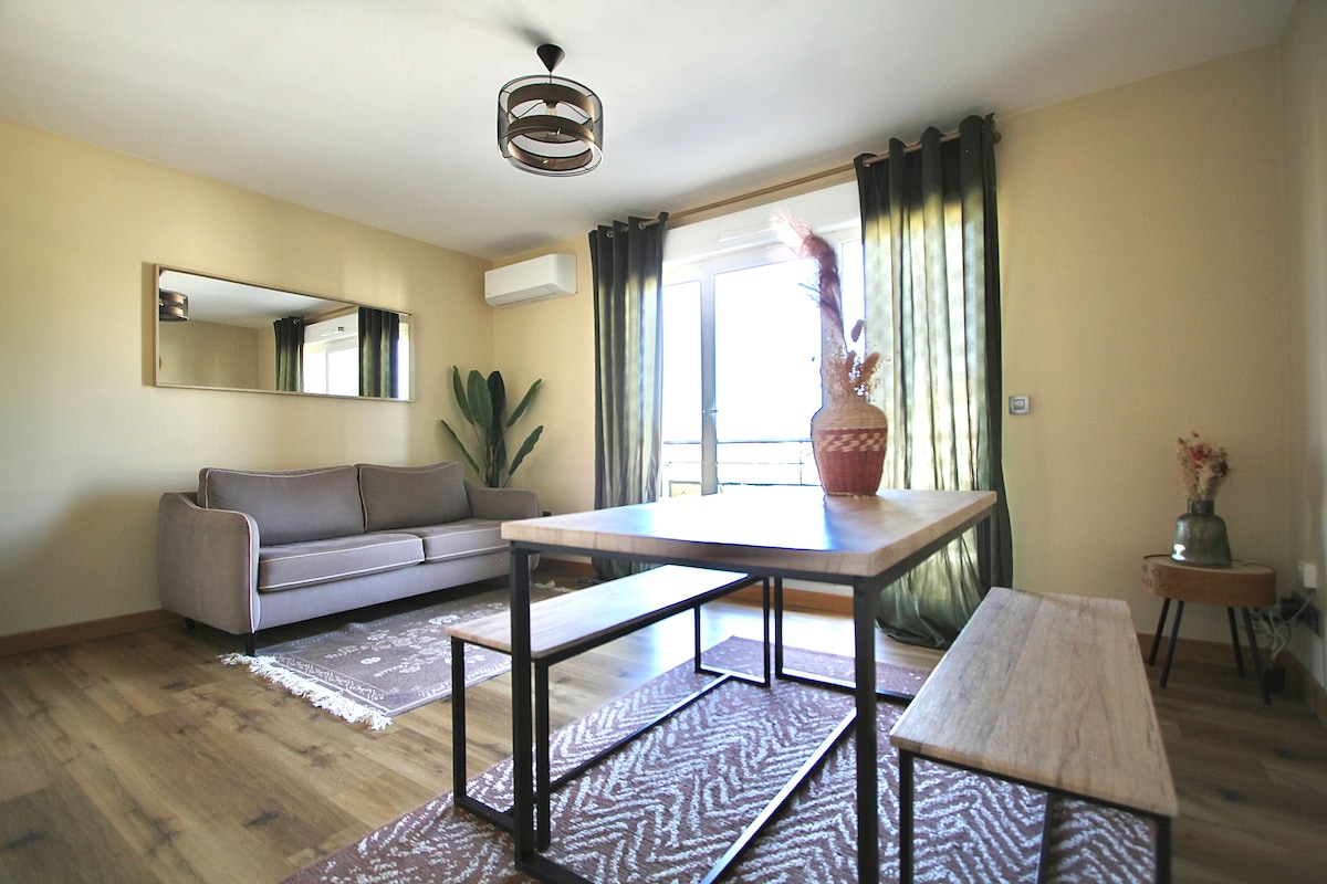 The living area features a soft sofa and a wooden dining table accompanied by benches, arranged on a textured rug. Floor-to-ceiling curtains frame the window, allowing natural light to illuminate the space. A large mirror reflects the room, enhancing the sense of openness.