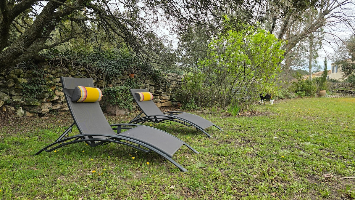 Two loungers with soft cushions are positioned on a lush green lawn, surrounded by trees and shrubs. A stone wall can be seen in the background, providing a natural backdrop to the serene garden space.