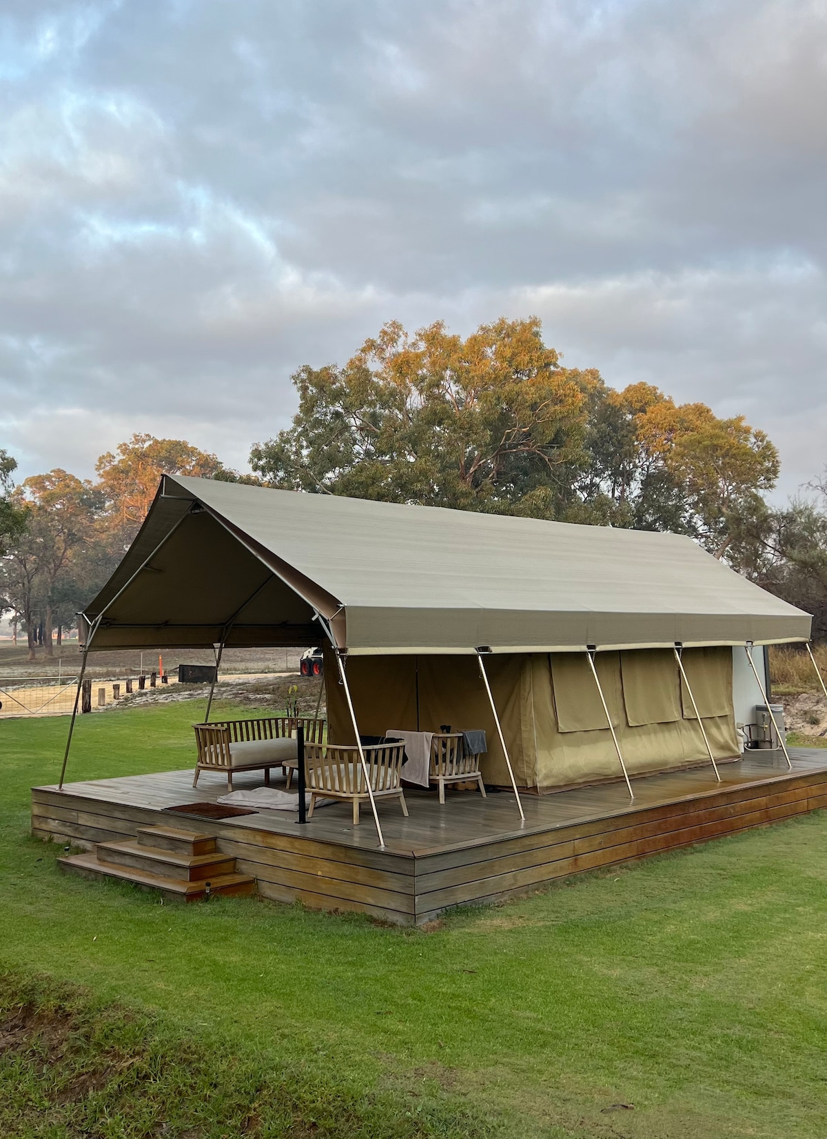 A spacious glamping tent is set on a wooden deck, surrounded by lush green grass. The tent features an awning entrance and is furnished with wooden chairs positioned around a table. Trees are visible in the background, providing a natural setting.