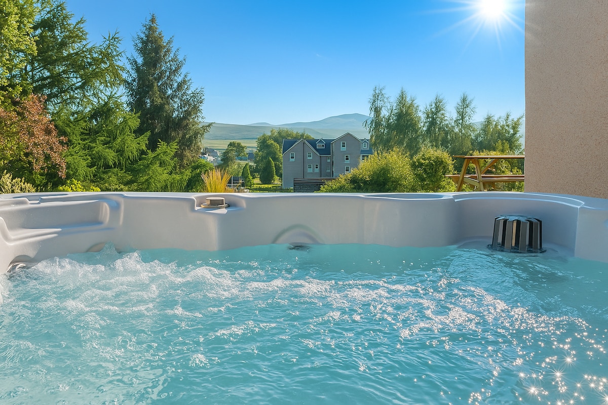 A hot tub filled with clear water is positioned with a view of surrounding greenery and distant hills under a bright blue sky. Sunlight reflects off the water, creating a serene atmosphere for relaxation.